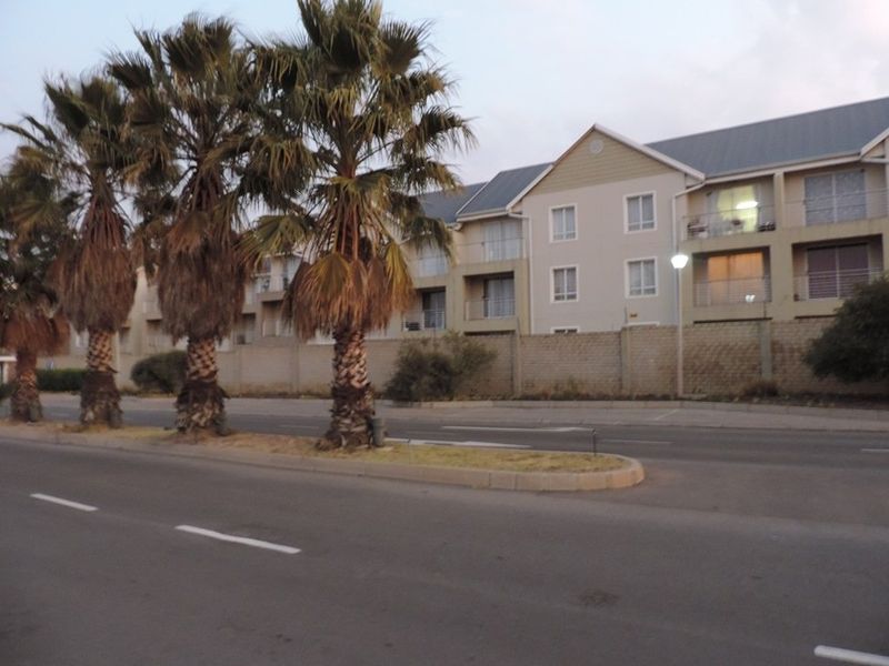 Street view of the apartment complex exterior, featuring a perimeter wall, mature palm trees, and paved road access.