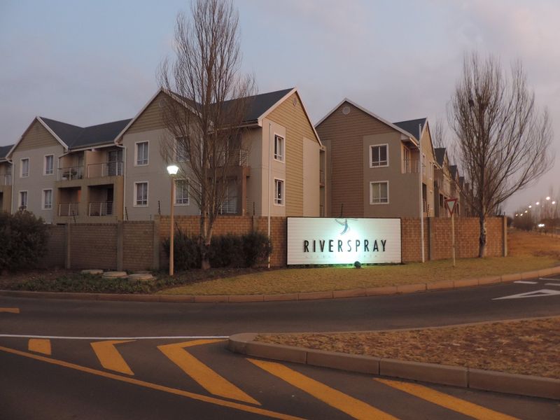 Street view of the Riverspray apartment complex entrance, featuring multi-story residential buildings, brick boundary walls, and integrated security fencing.