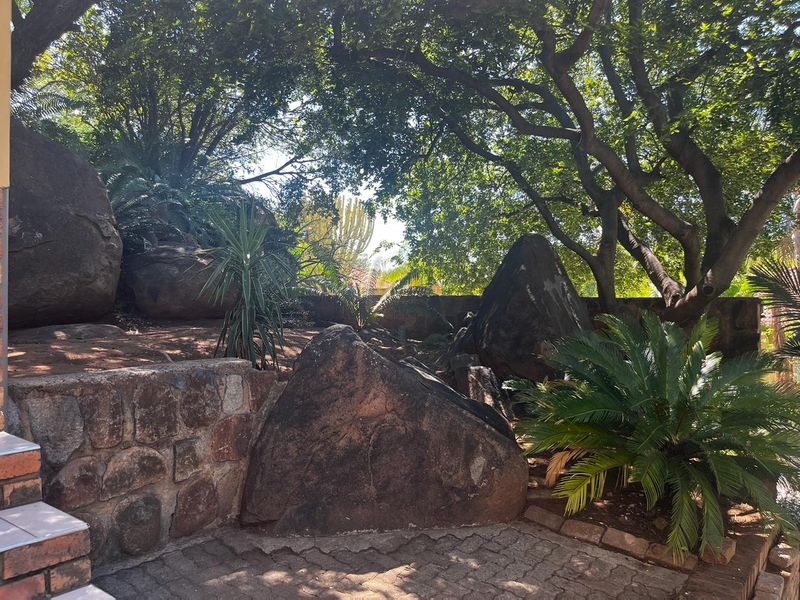 A landscaped garden area featuring mature trees, large natural boulders, and a stone retaining wall with paved ground surfacing.