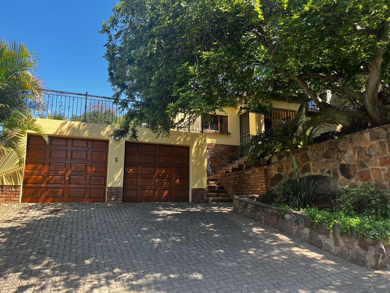 Front elevation featuring a double garage, paved driveway, stone retaining wall, and an upper-level balcony with wrought iron railings.