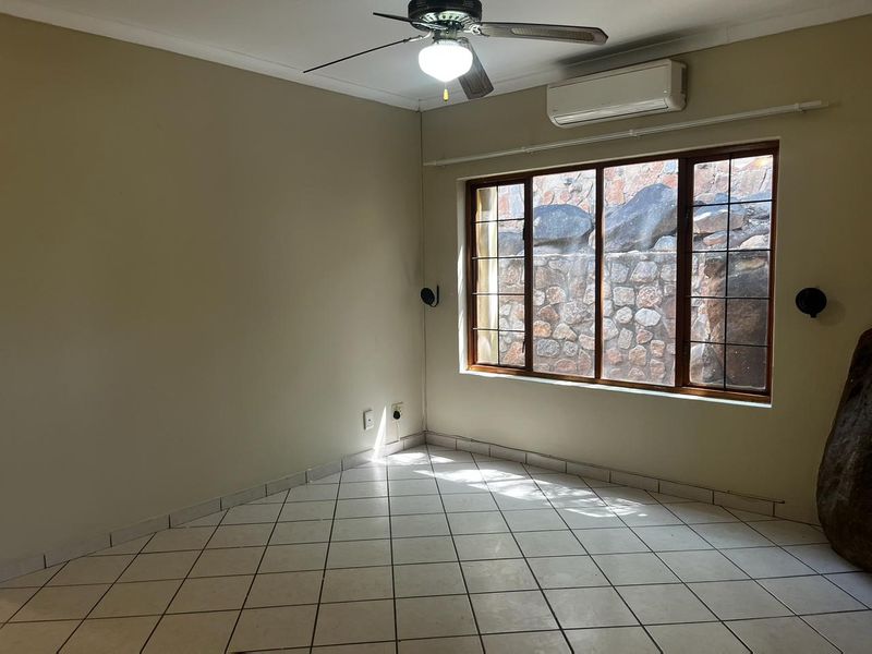 A spacious, empty bedroom featuring tiled flooring, a ceiling fan, and a wall-mounted air conditioning unit, with a view of a stone retaining wall.