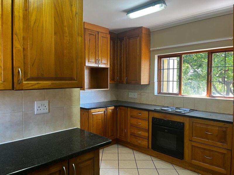 A functional kitchen featuring wooden cabinetry, granite countertops, a built-in oven, and an electric hob, with tiled flooring and a window overlooking the garden.