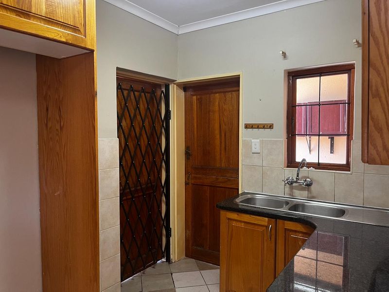 A functional scullery area featuring wooden cabinetry, granite countertops, a double stainless steel sink, and direct access to an exterior door with a security gate.