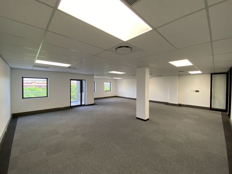 A spacious, open-plan commercial office area featuring neutral carpet tiles, a suspended ceiling with recessed lighting, and large windows providing natural light.