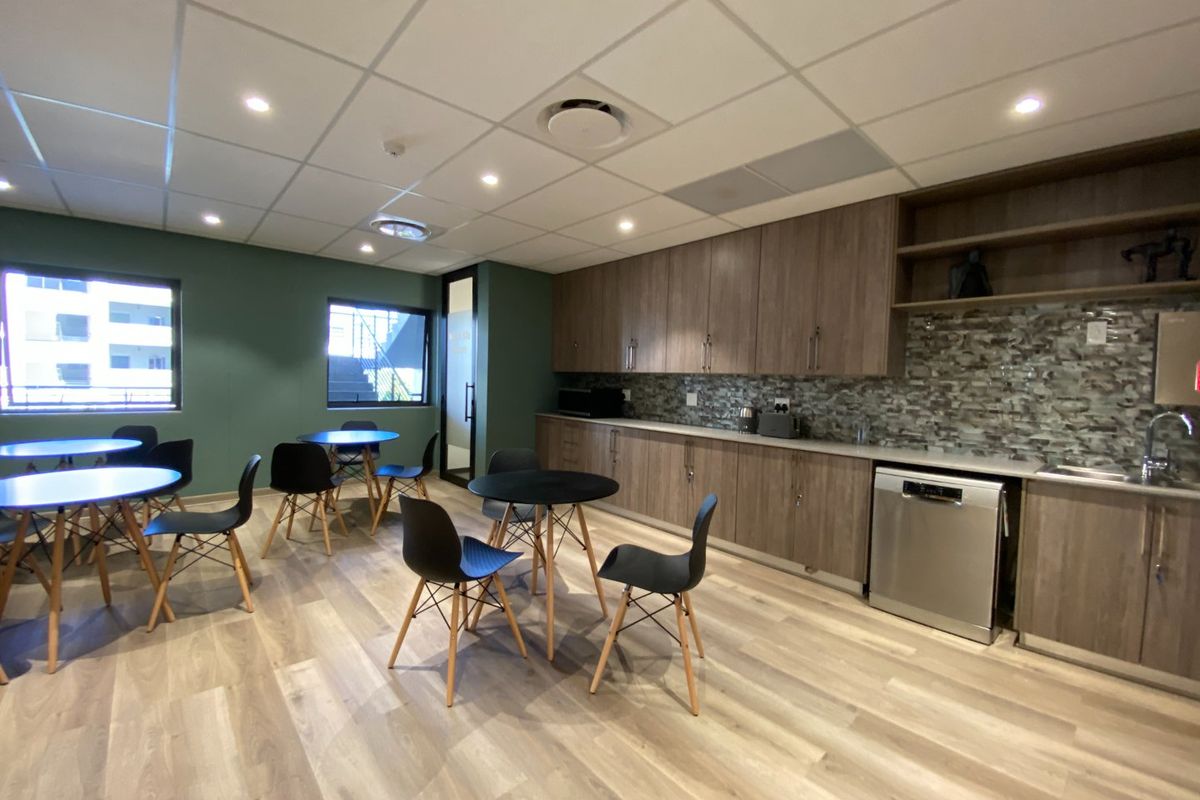 A modern commercial kitchenette featuring wood-finish cabinetry, mosaic tile backsplash, integrated dishwasher, and durable laminate flooring in a bright, open-plan office break area.