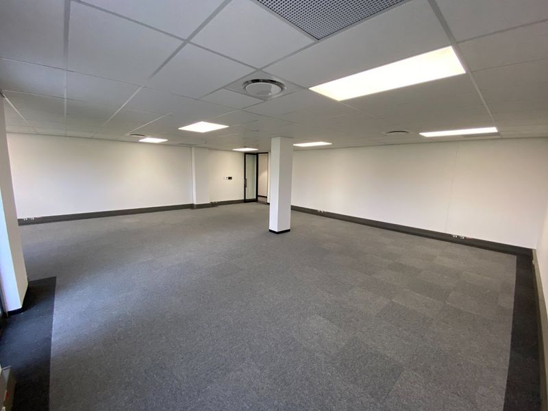 A spacious, empty commercial office area featuring neutral carpet tiles, a suspended ceiling with recessed lighting, and a central structural pillar.