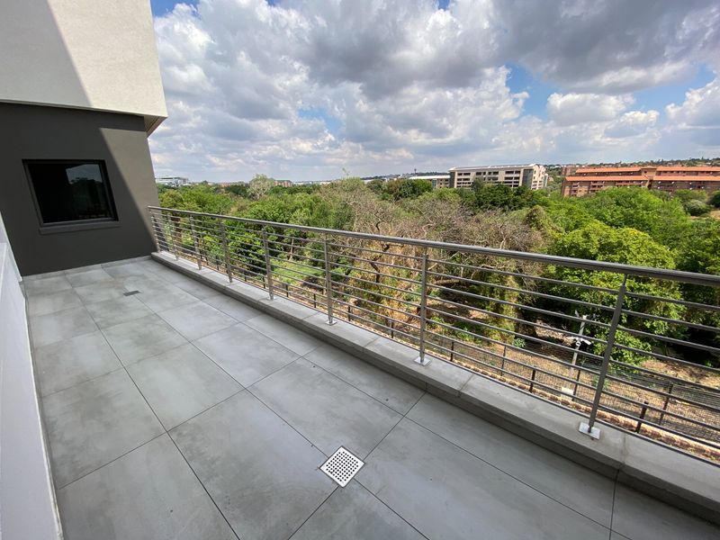 A modern, spacious balcony featuring tiled flooring and a stainless steel balustrade, offering an elevated view of the surrounding greenery and urban landscape.