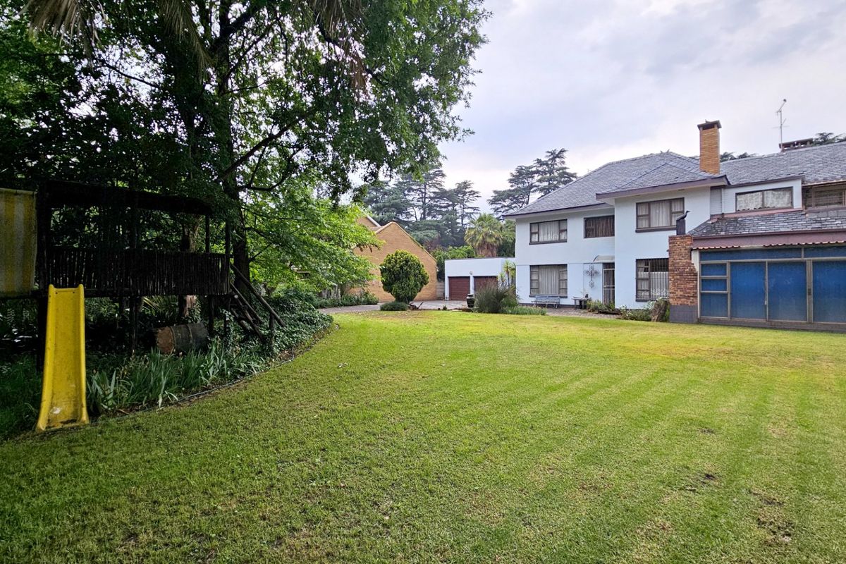 A spacious rear garden view featuring a large lawn, mature trees, and the rear elevation of a multi-story residential house with a chimney.