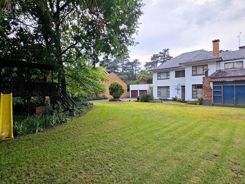 A spacious rear garden view featuring a large lawn, mature trees, and the rear elevation of a multi-story residential house with a chimney.