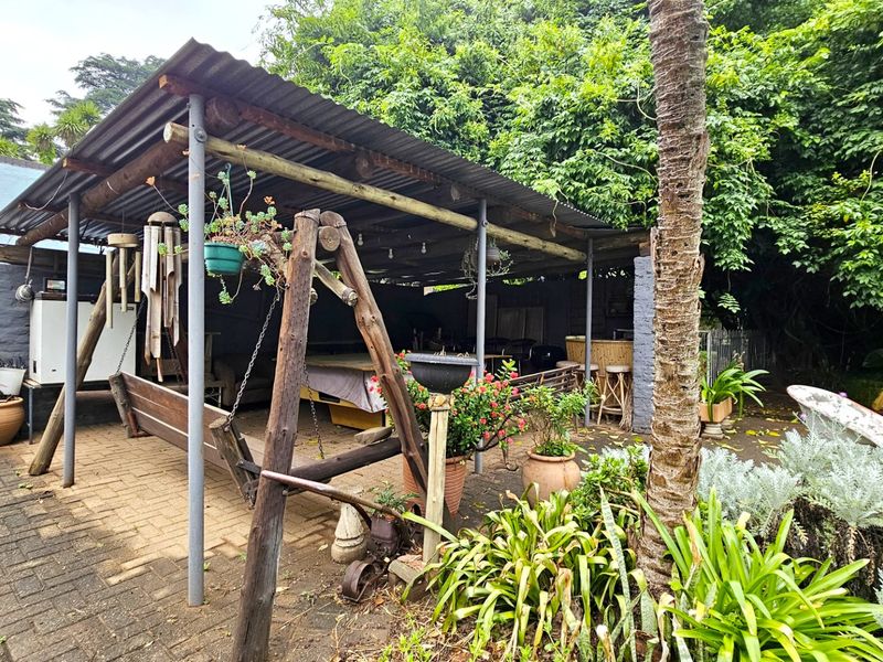 A covered outdoor patio area featuring a corrugated iron roof, timber support structure, and paved flooring, set within a lush garden environment.