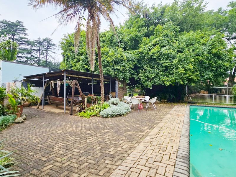 Outdoor pool area featuring a paved patio and a covered entertainment structure, surrounded by mature garden landscaping.