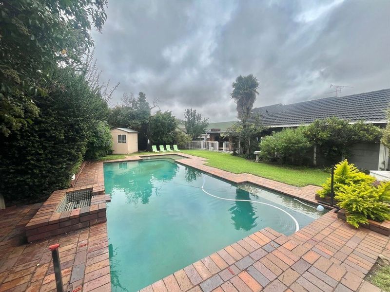 A backyard area featuring a swimming pool with brick paving, a small garden shed, and established lawn and garden beds.