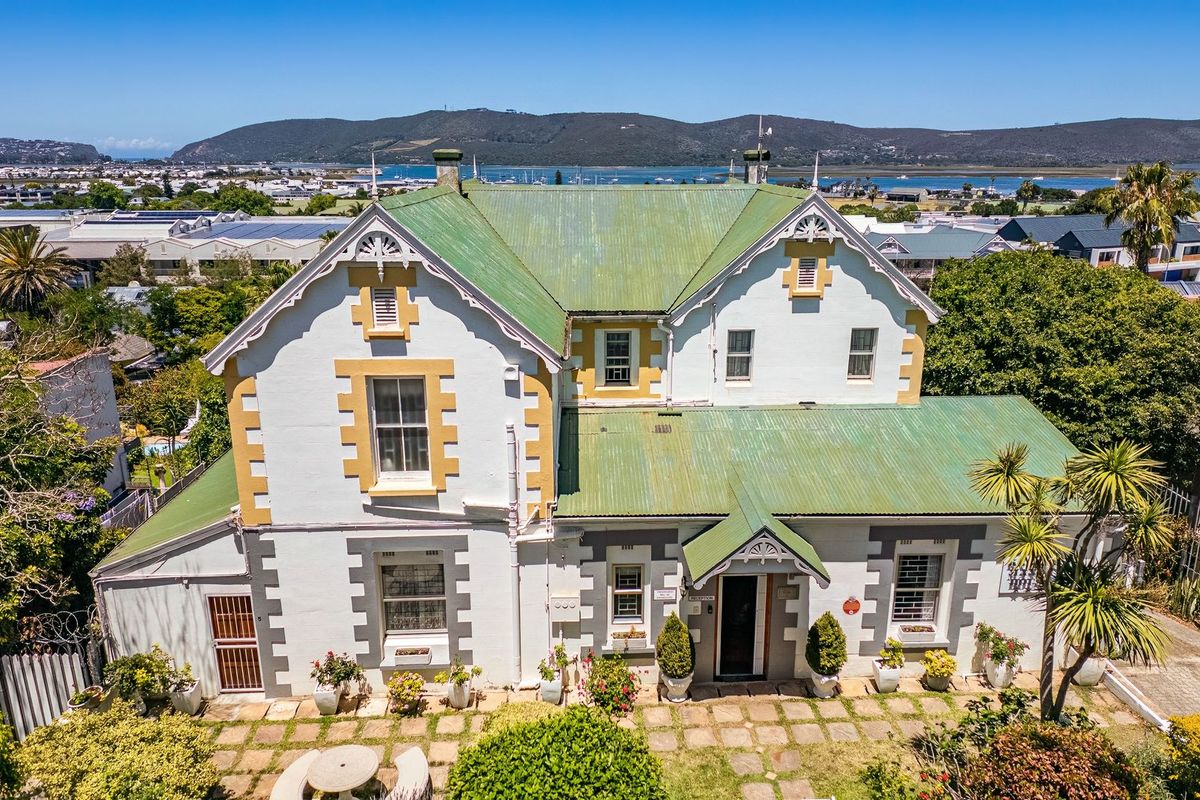 Aerial view of a historic-style commercial property featuring a green corrugated metal roof, stone-paved patio area, and scenic coastal views in the background.