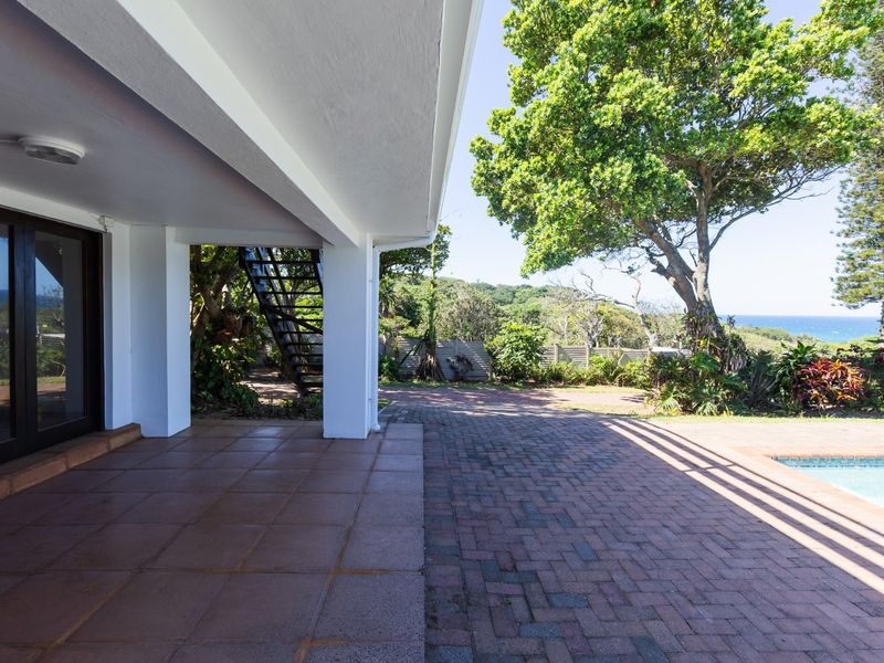 A spacious covered patio area featuring brick paving, overlooking a swimming pool and garden with ocean views in the distance.