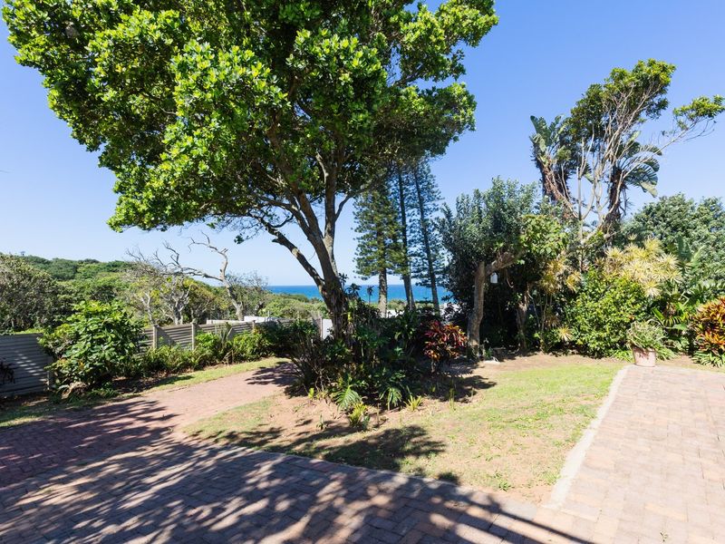 A well-maintained garden area featuring mature trees, paved pathways, and boundary fencing with a distant ocean view.