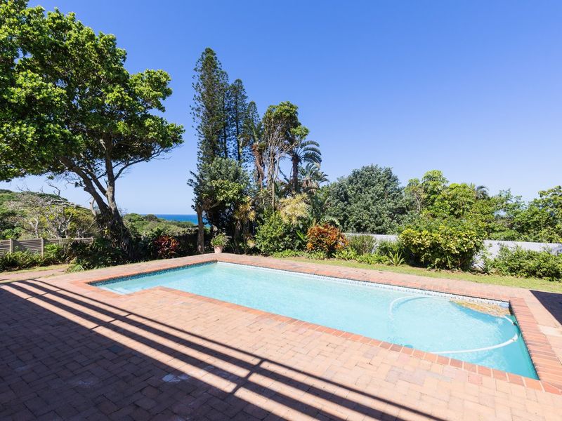 A well-maintained swimming pool area featuring brick paving, lush garden surroundings, and a partial view of the ocean in the distance.