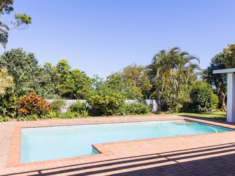 A well-maintained swimming pool area featuring brick paving and a covered patio structure, set within a lush, established garden.