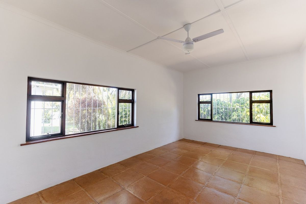 A bright, empty bedroom featuring tiled flooring, a ceiling fan, and large windows fitted with security bars overlooking the garden.