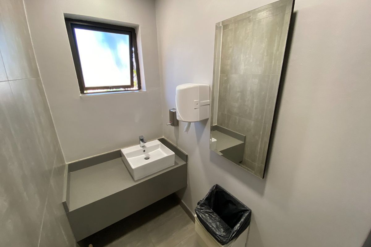 A clean, modern commercial restroom featuring a wall-mounted basin, tiled vanity, and wall-mounted mirror with neutral finishes.