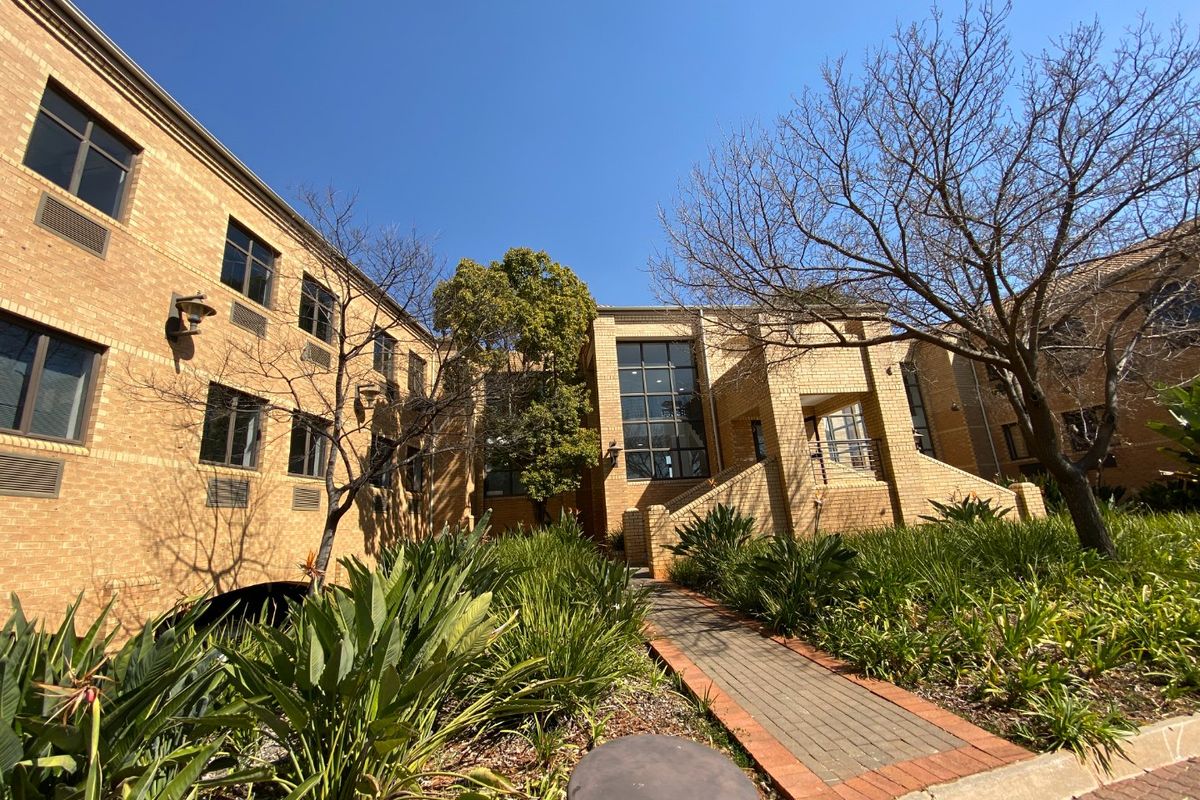 A well-maintained commercial building exterior featuring face brick construction, large glass windows, and a paved walkway leading through landscaped garden areas.