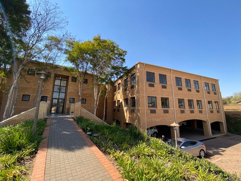 A well-maintained commercial building featuring a brick facade, paved pedestrian walkway, and integrated covered parking area with landscaped surroundings.