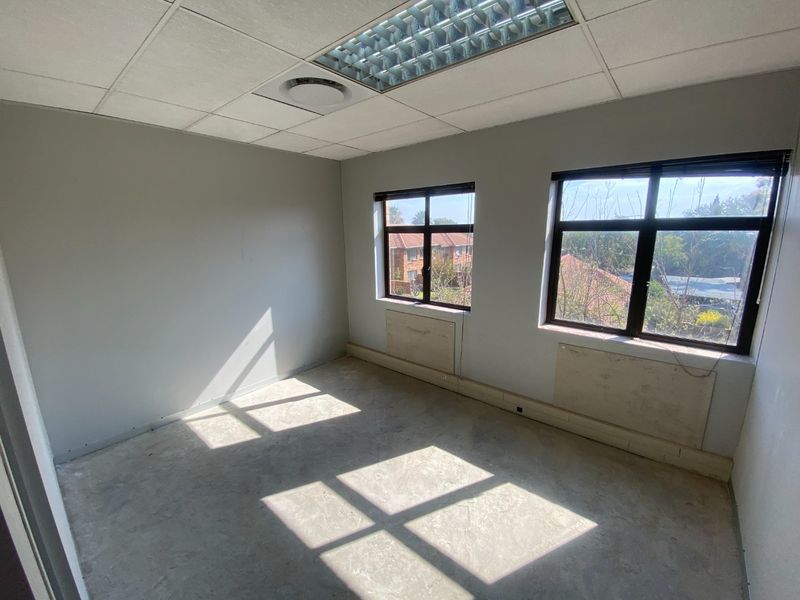An empty commercial office space featuring a suspended ceiling, recessed lighting, and large windows with natural light, currently requiring floor and wall finishing.