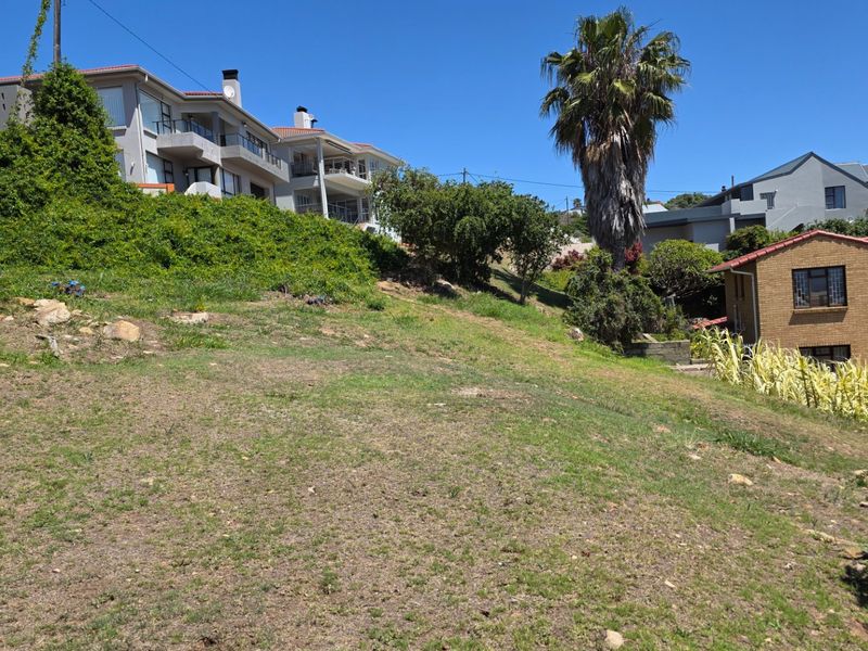 A view of a sloped, vacant residential plot with surrounding established neighborhood properties.
