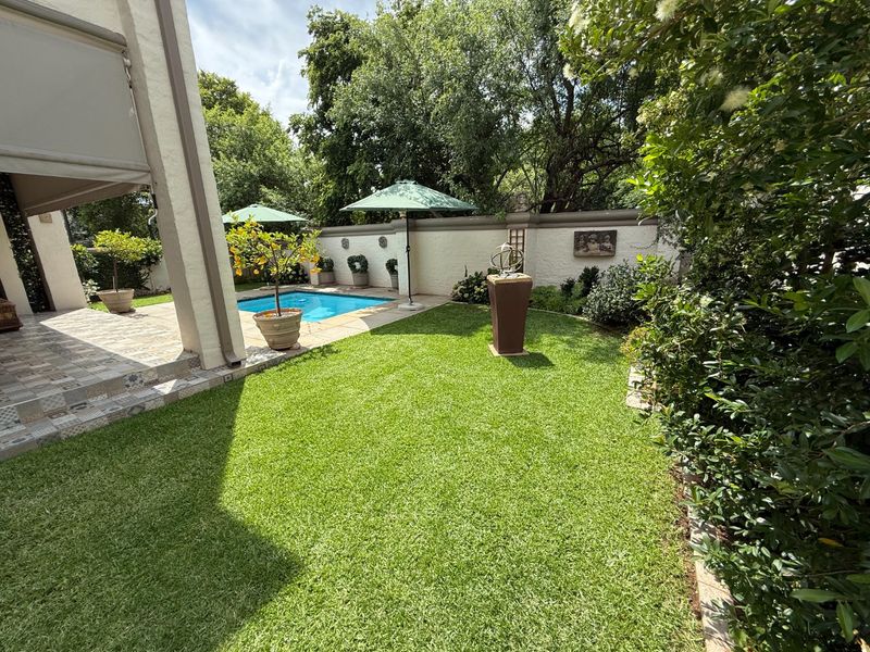 A well-maintained outdoor area featuring a swimming pool, a tiled patio space, and a lush green garden enclosed by a boundary wall.
