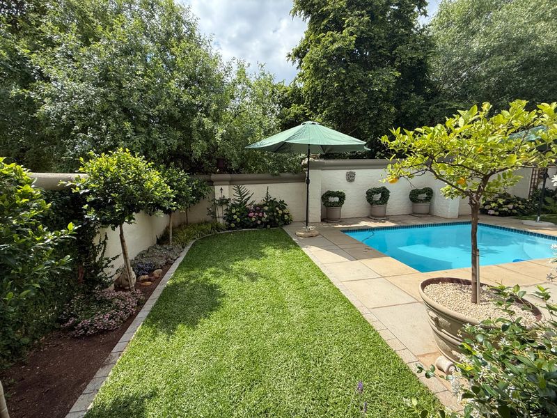 A well-maintained outdoor pool area featuring a clear swimming pool, paved surround, and established garden landscaping with a boundary wall.
