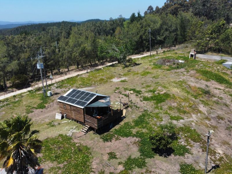 Aerial view of a rural property featuring a small timber cabin with solar panels, a deck, and an outdoor fireplace set on a large plot.
