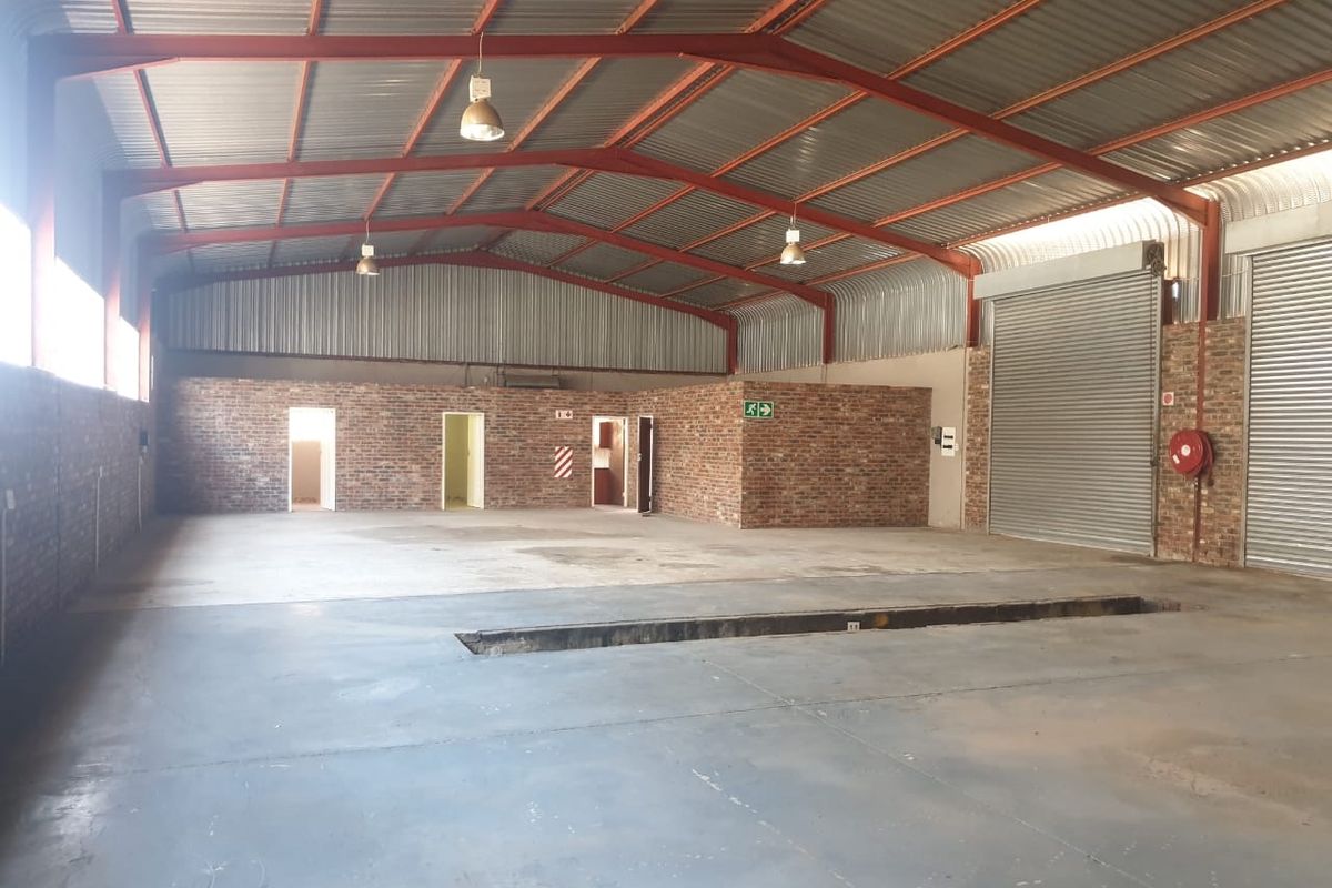 Spacious industrial warehouse featuring exposed brick walls, high-clearance corrugated metal roofing, concrete flooring, roller shutter doors, and a built-in vehicle inspection pit.