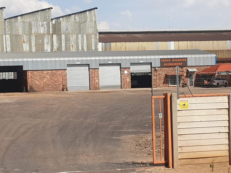 Industrial facility exterior featuring multiple roller shutter doors, brick facade, and corrugated metal roofing, secured by a perimeter gate.