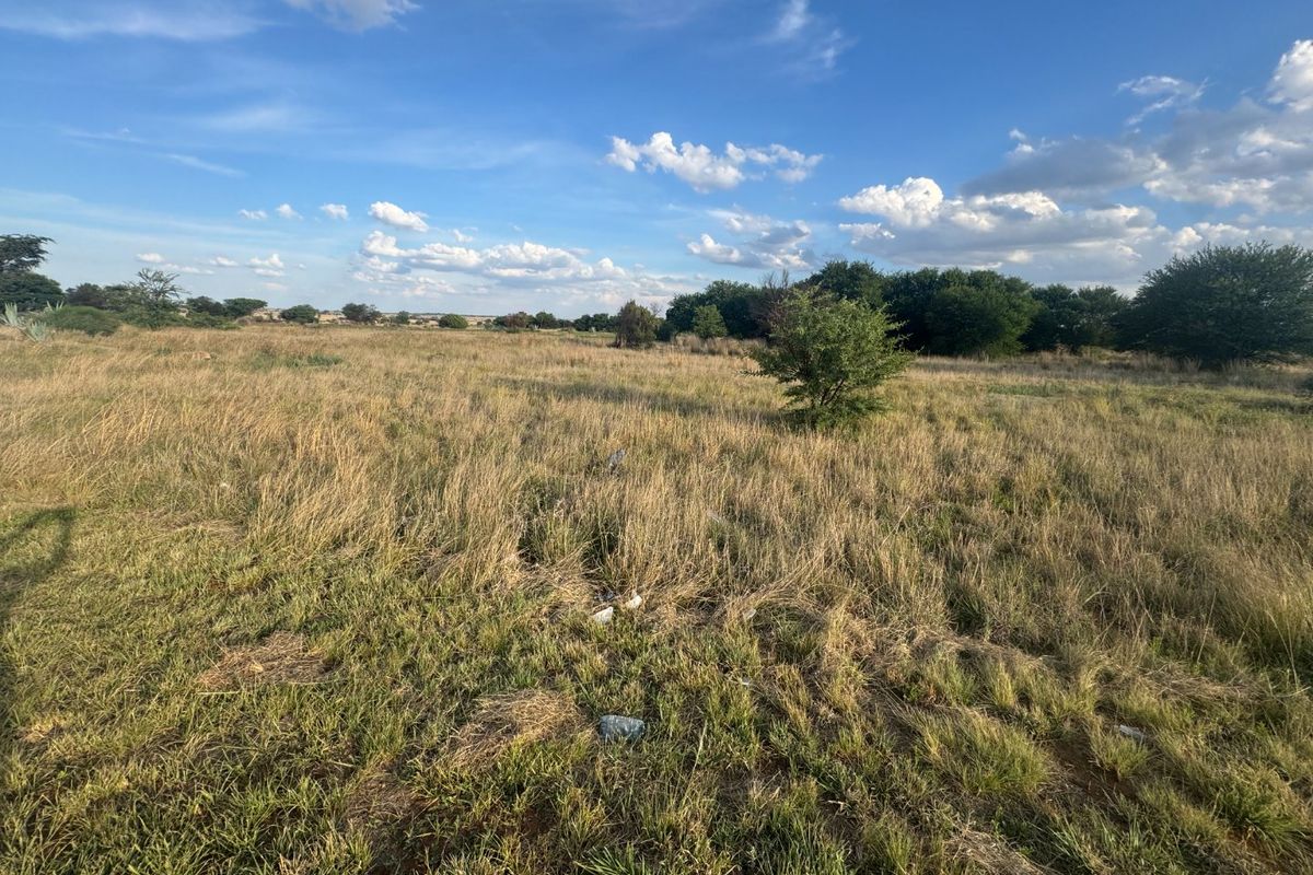 A wide-angle view of an undeveloped residential vacant land plot featuring natural grass and scattered shrubs under a clear blue sky.