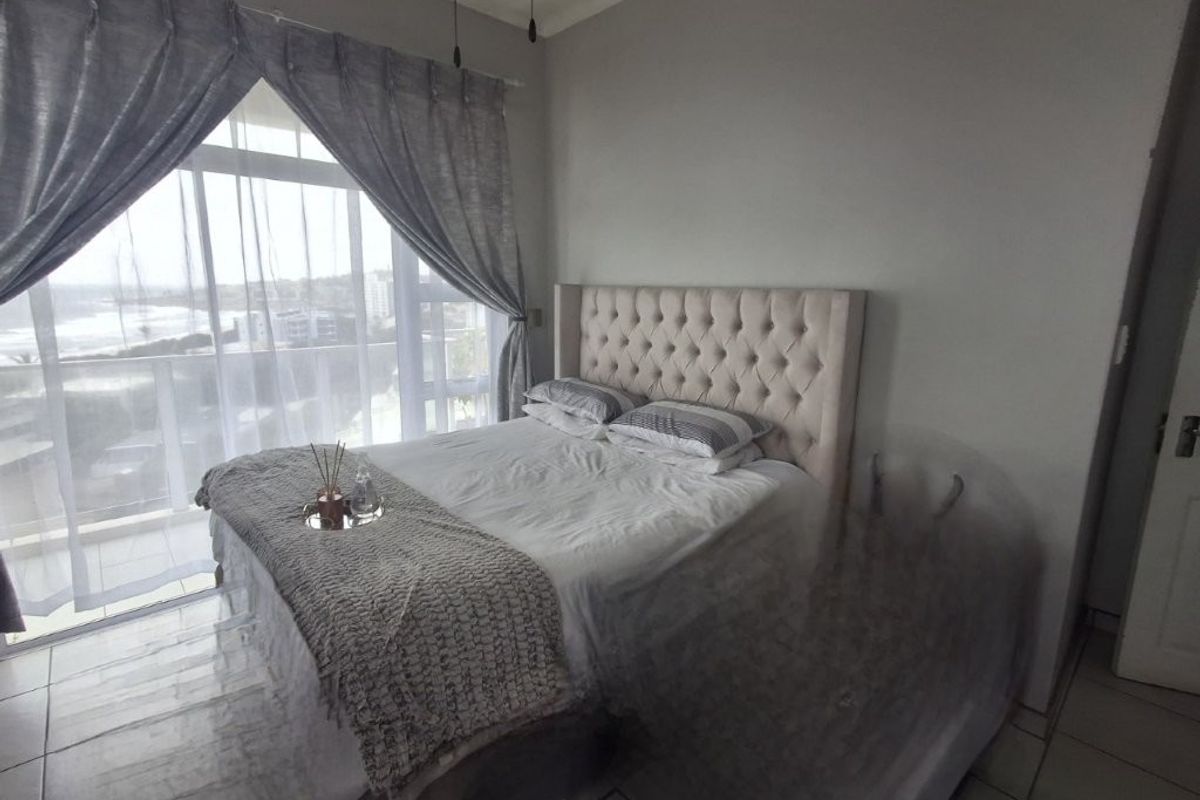 A bright bedroom featuring large sliding glass doors that open onto a balcony with coastal views, tiled flooring, and neutral wall finishes.