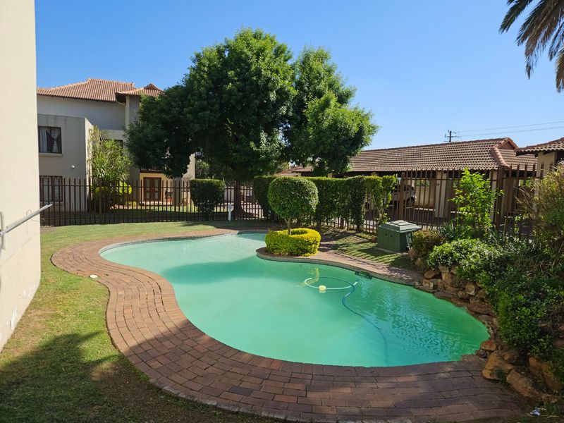 A well-maintained outdoor swimming pool area featuring brick coping, established garden landscaping, and secure metal perimeter fencing.
