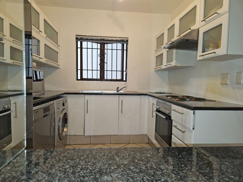 Compact kitchen featuring white cabinetry, granite countertops, built-in oven, electric hob, and integrated laundry space with security bars on the window.