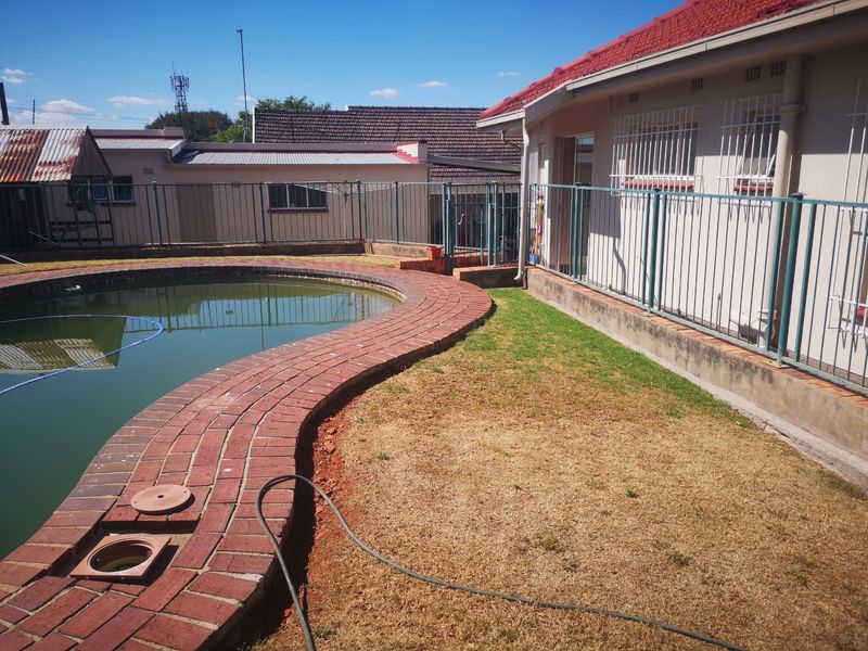 Rear exterior view featuring a swimming pool with brick coping, metal security fencing, and a lawn area adjacent to the main house structure.