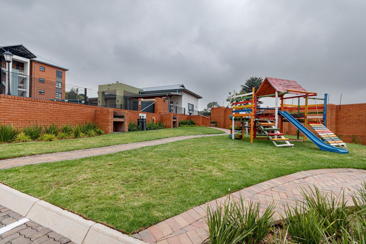 A well-maintained communal garden area featuring a paved walkway, built-in brick braai facilities, and a playground set within a secure complex.