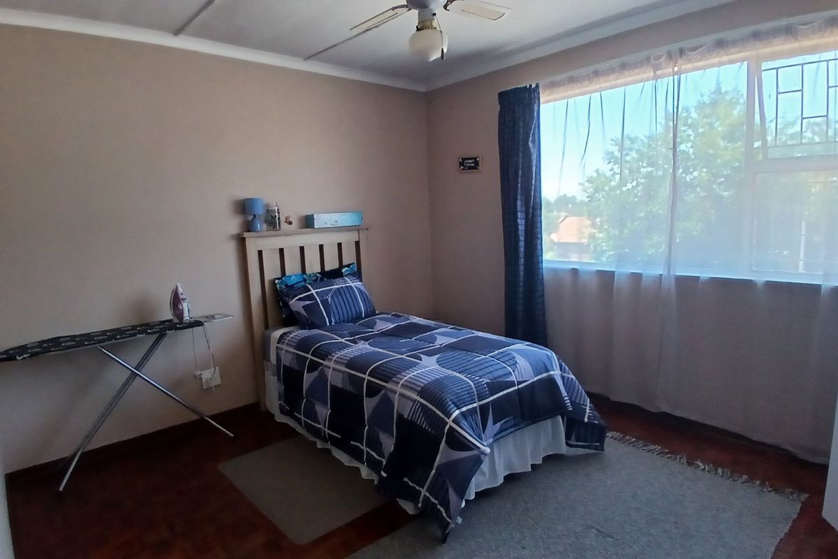 A functional bedroom featuring wooden flooring, a ceiling fan, and a large window with security bars, providing natural light.