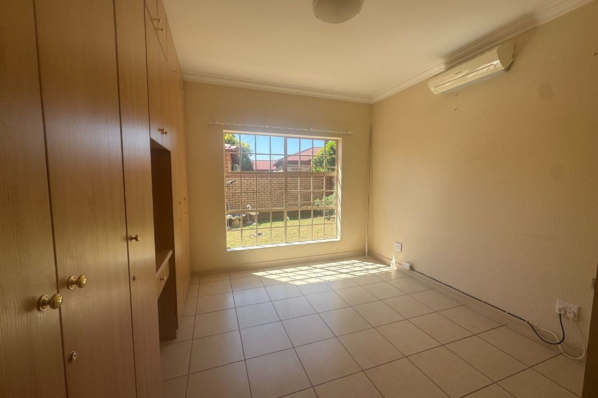 A bright, empty bedroom featuring tiled flooring, built-in wooden cupboards, and a wall-mounted air conditioning unit with a view of the rear garden.