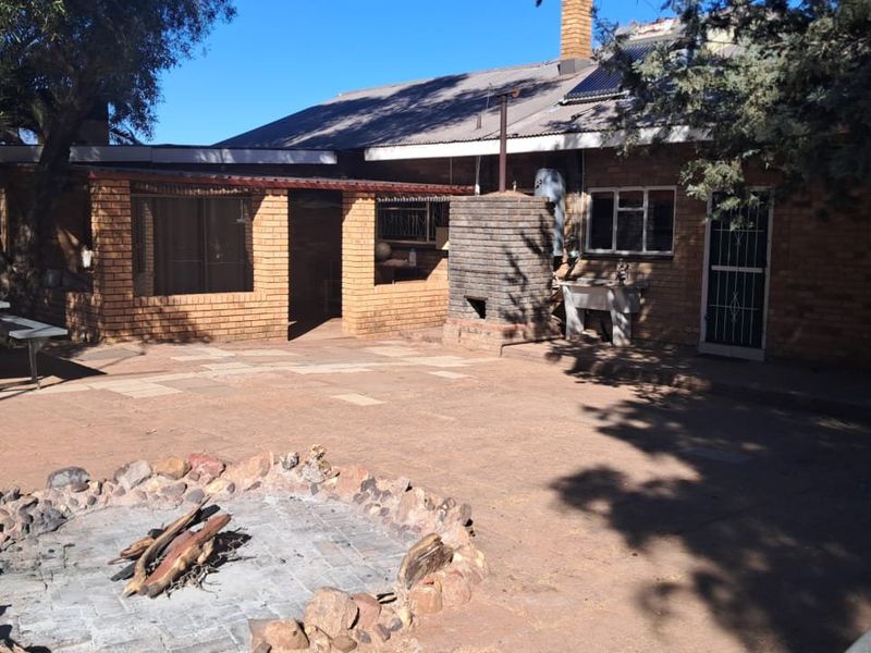 Rear exterior view of a brick farmhouse featuring a paved patio, built-in braai area, and a stone fire pit in the garden.