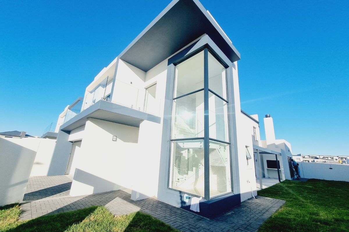 Modern double-story residence featuring a striking double-height glass facade, paved outdoor area, and integrated built-in braai, showcasing contemporary architectural design.