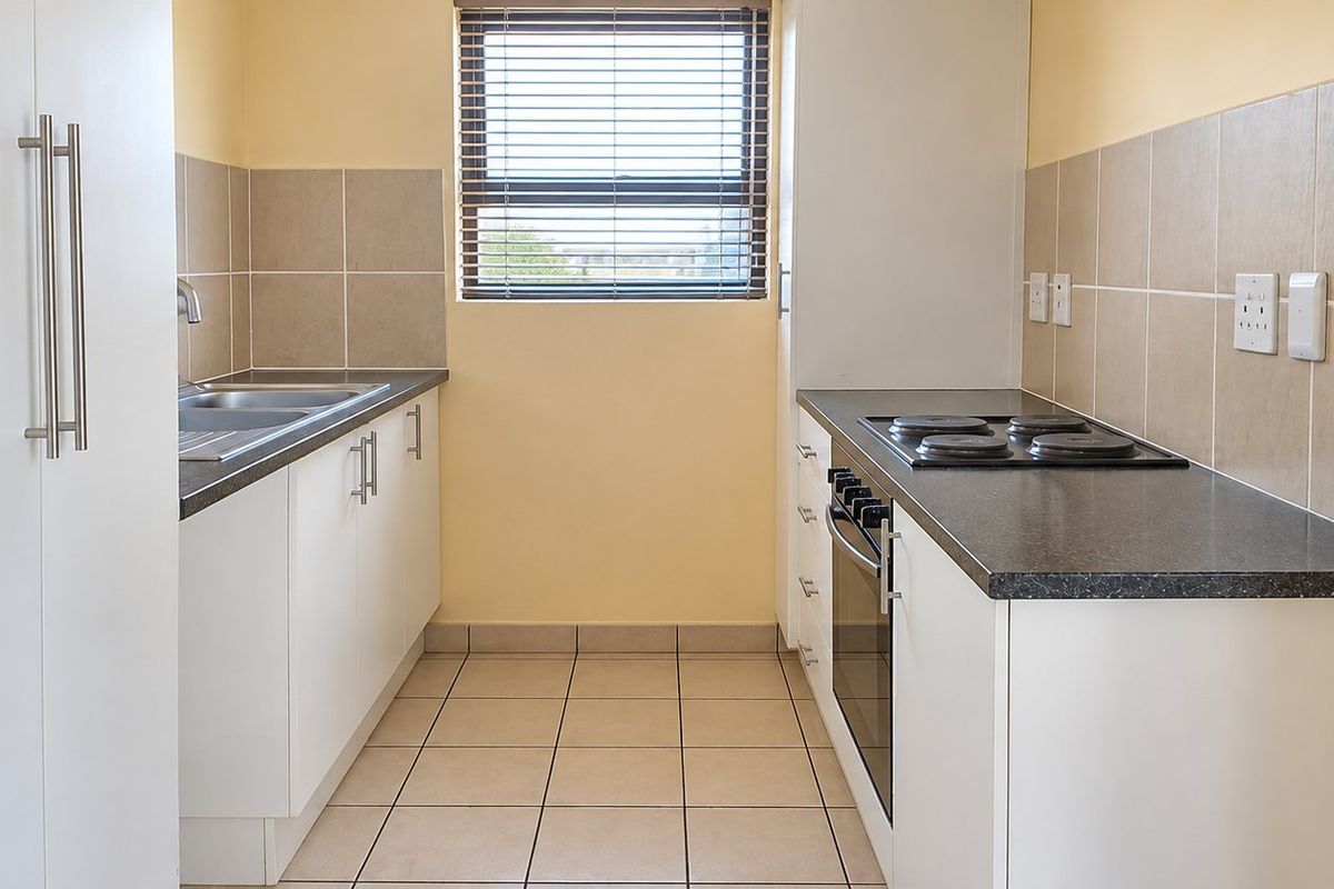 A clean, functional galley-style kitchen featuring white cabinetry, dark laminate countertops, a built-in oven, electric hob, and neutral tiled splashbacks.