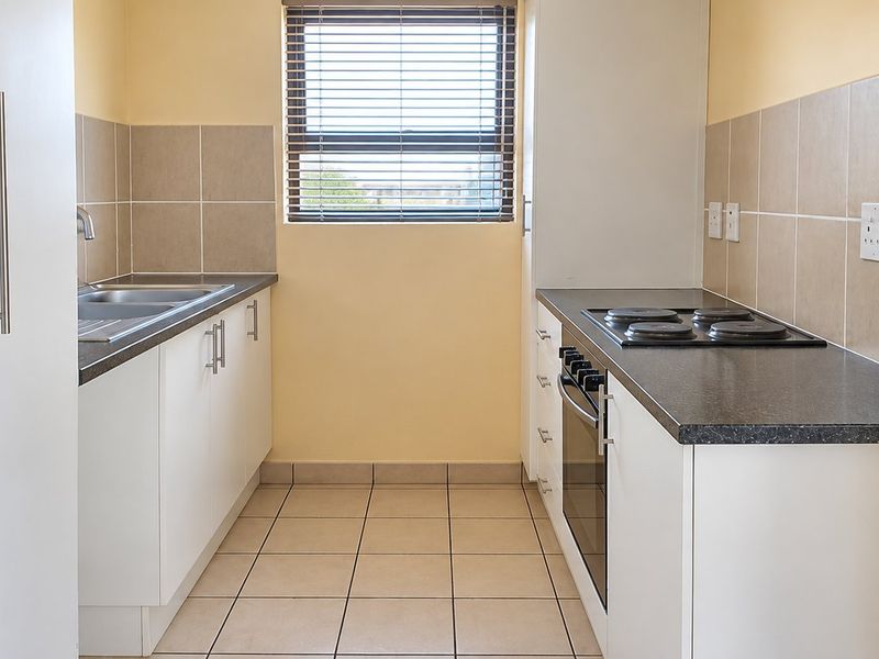 A clean, functional galley-style kitchen featuring white cabinetry, dark laminate countertops, a built-in oven, electric hob, and neutral tiled splashbacks.