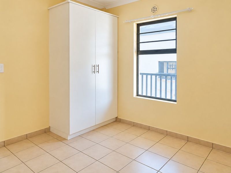 A clean, empty bedroom featuring neutral yellow walls, tiled flooring, and a built-in white wardrobe with a window providing natural light.
