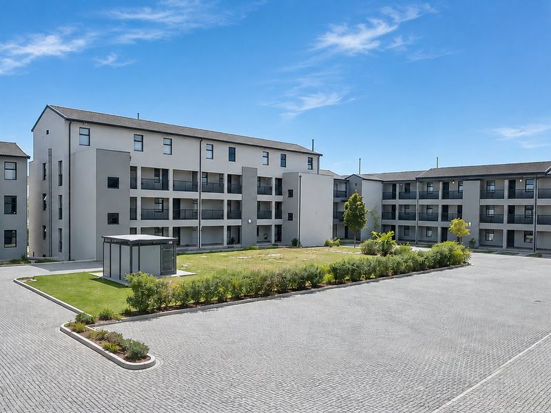 A wide-angle view of the apartment complex exterior, featuring paved parking areas, communal garden spaces, and multi-story residential blocks with balconies.