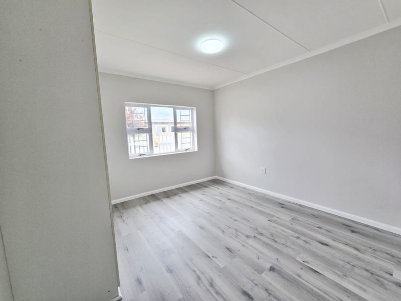 A bright, empty bedroom featuring modern grey laminate flooring, neutral walls, and a window fitted with security bars.