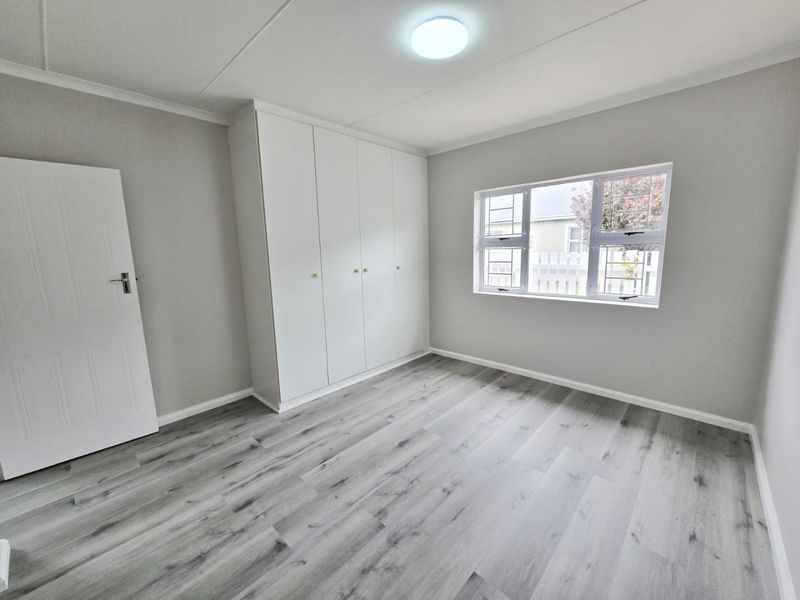 A bright, empty bedroom featuring modern grey laminate flooring, neutral walls, and white built-in cupboards with security bars on the window.
