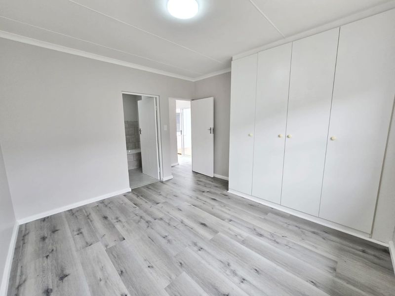 A bright, empty bedroom featuring modern grey laminate flooring, neutral walls, and ample built-in cupboard storage with an en-suite bathroom access.