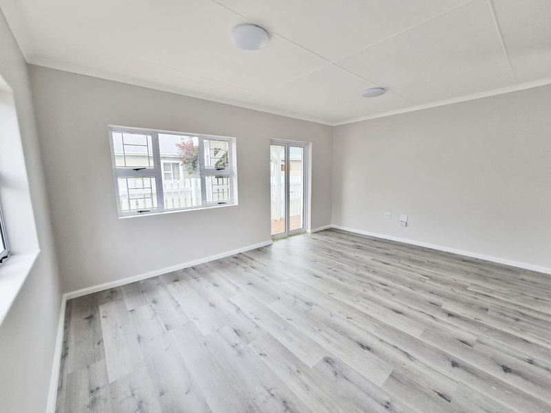 A bright, empty living room featuring modern grey laminate flooring, neutral walls, and access to an outdoor area through a glass sliding door.